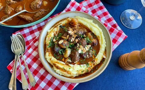 A bowl of braised stew with creamy mash on a red table cloth, topped with almonds and herbs
